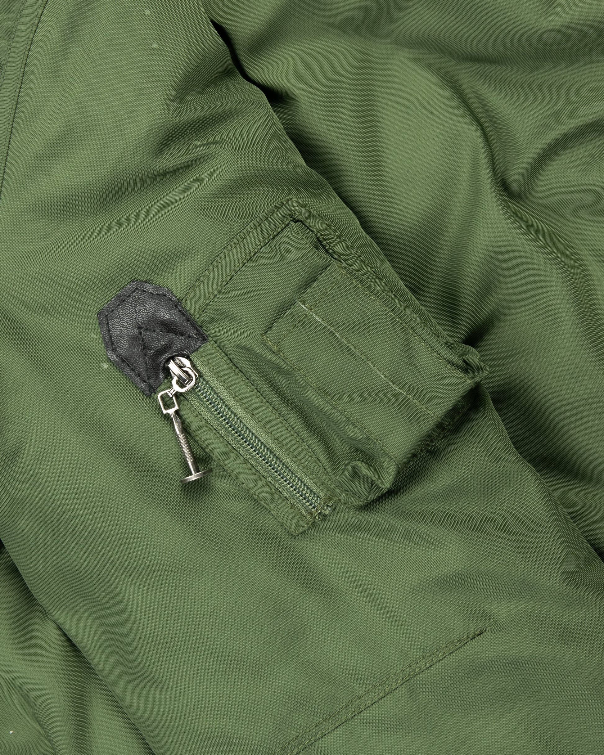 Close-up of men’s khaki green jacket sleeve featuring a zippered pocket with metal pull tab and stitched detail, highlighting functional design.